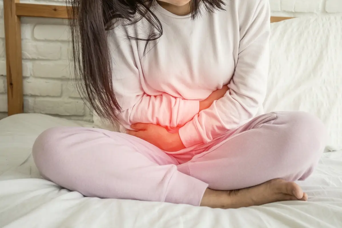 A woman sitting on a bed holding her stomach, with the uncomfortable area marked in red